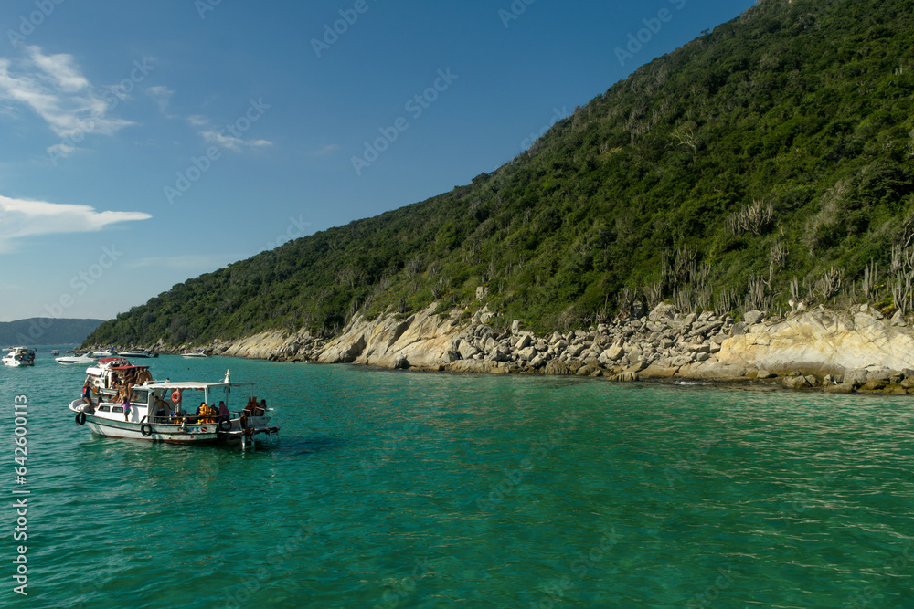 Formação rochosa a beira-ma com muita vegetação, céu azul e pequena embarcação no mar, localizada na região de Cabo Frio, Rio de Janeiro, Brasil - 30