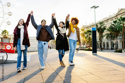 Happy group of young people having fun together walking in city street. Joyful diverse friends holding hands celebrating outdoors. Youth, friendship and travel concept.