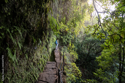 Female backpacker tourist walking along rainforest water channel path on steep cliff covered with plants. Levada of Caldeirão Verde, Madeira Island, Portugal, Europe.