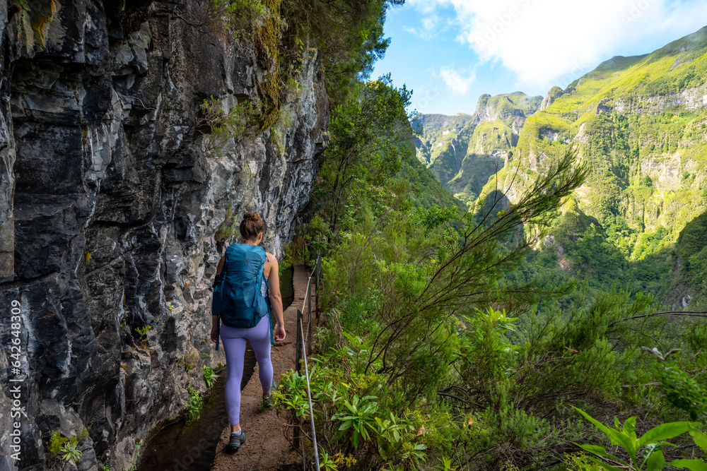 Backpacker walks along a picturesque hike trail under a large steep rock face along a water channel through Madeira rainforest. Levada of Caldeirão Verde, Madeira Island, Portugal, Europe.