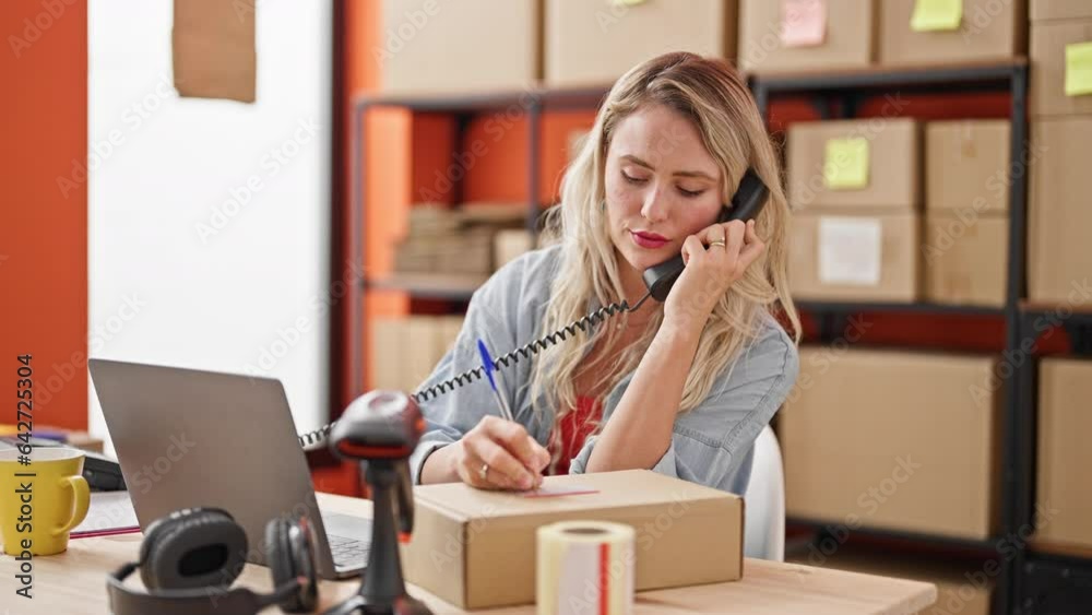 Young blonde woman ecommerce business worker talking on telephone writing on package at office Young blonde woman ecommerce business worker talking on telephone writing on package at office