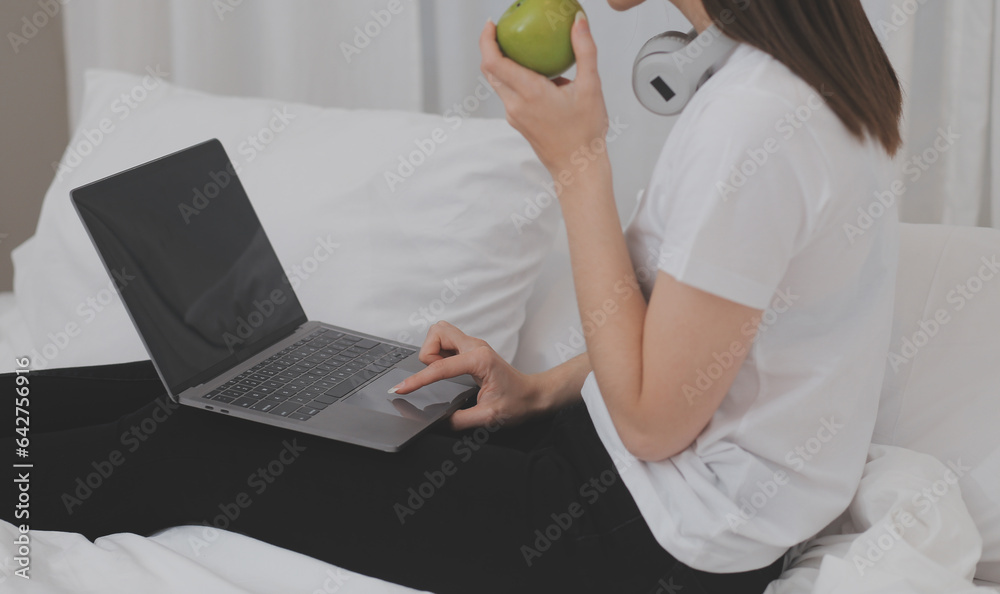 Naklejka premium Efficient Young Millennial Girl Sitting on a Bed in the Morning, Uses Laptop Computer and Eats Croissants and Drinks Coffee for Breakfast