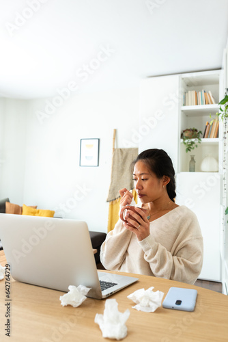Chinese woman feel sick working from home. Asian female blowing nose with tissue holding cup of herbal tea. Vertical