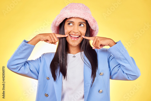 Fashion, smile and face of Indian woman on yellow background with silly, goofy and playful expression. Emoji, happiness and person in studio pointing to mouth in trendy accessories, style and hat