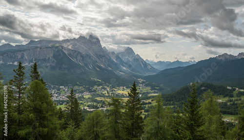 View on the surrounding mountains of Cortina D'Ampezzo in Italy from Tofana mountain. Pine trees in the foreground, mountain range in the background. Cloudy summer day.