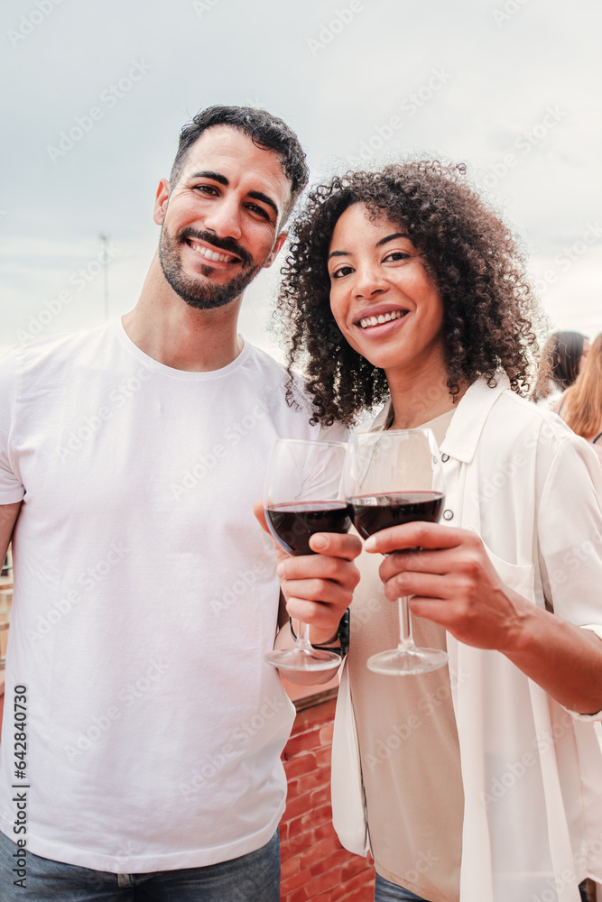 Vertical portrait of a multiracial couple toasting the wine glasses, smiling and looking at camera together, clinking on rooftoop a party celebration. Patio social reunion at home. High quality photo