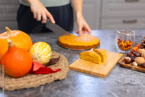 A woman cuts a cake on the kitchen table and puts it on a wooden board. Delicious baked dessert made from flour and pumpkin. Orange pie. Pie, pumpkins and nuts. Autumn dessert