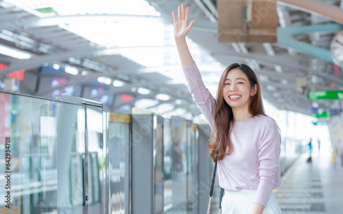 Asian woman smiling with happiness and refresh, wearing casual business shirt, waiving hand to greet, meet friend as appointment, standing on platform, waiting for sky train transportation.