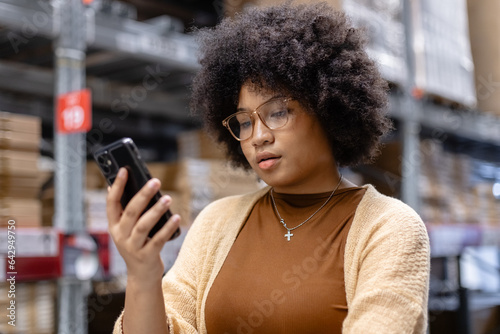 Businesswoman African American afro hairstyle using smartphone standing in warehouse. Customers look for products on vacant shelf. woman worker check stock inspecting in logistic factory.