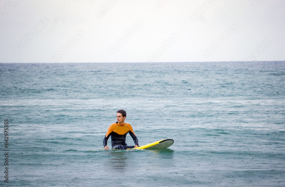 20s Male Surfer in Orange Neoprene Sitting on His Yellow Board Waiting to Catch a Good Wave in the Sea