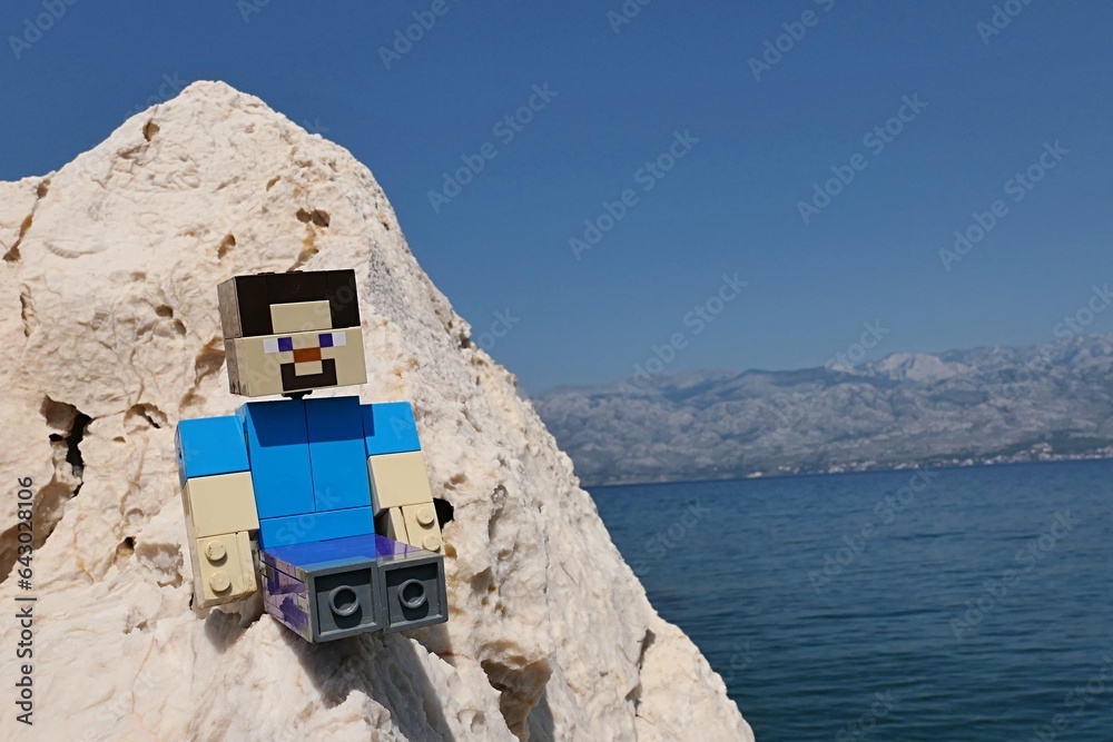 Naklejka premium LEGO Minecraft large figure of main character Steve sitting on rocky cliff of Radanovac beach near Razanac, northern Dalmatia, Croatia. Velebit mountain in background, august bright sunshine.