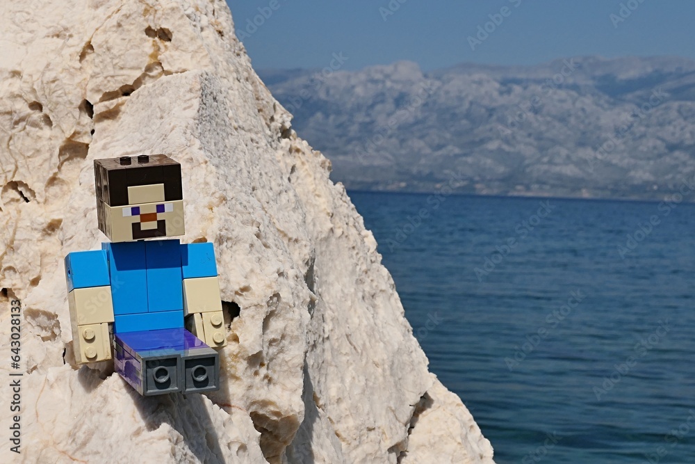 Naklejka premium LEGO Minecraft large figure of main character Steve sitting on rocks of Radanovac beach near Razanac, northern Dalmatia, Croatia. Velebit mountain in background, august bright sunshine.