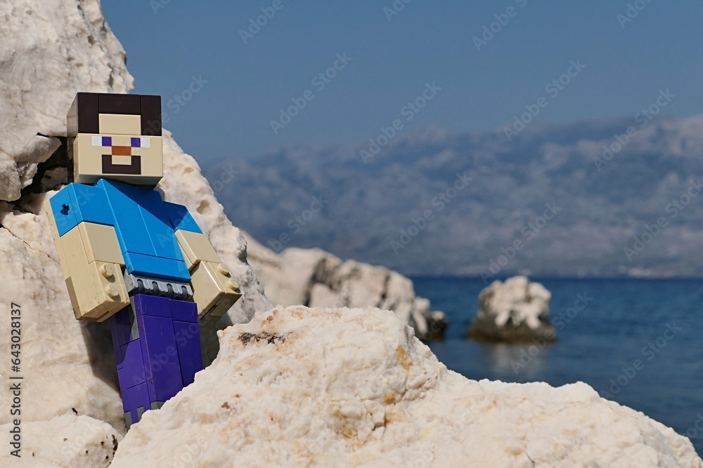 Naklejka premium LEGO Minecraft large figure of main character Steve posing on rocks of Radanovac beach near Razanac, northern Dalmatia, Croatia. Velebit mountain in background, august bright sunshine.
