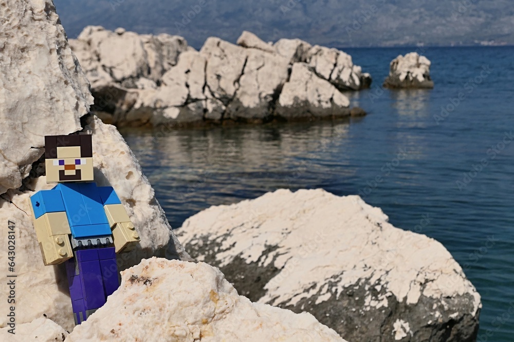 Naklejka premium LEGO Minecraft large figure of main character Steve standing on rocks of Radanovac beach near Razanac, northern Dalmatia, Croatia. Velebit mountain in background, august bright sunshine.