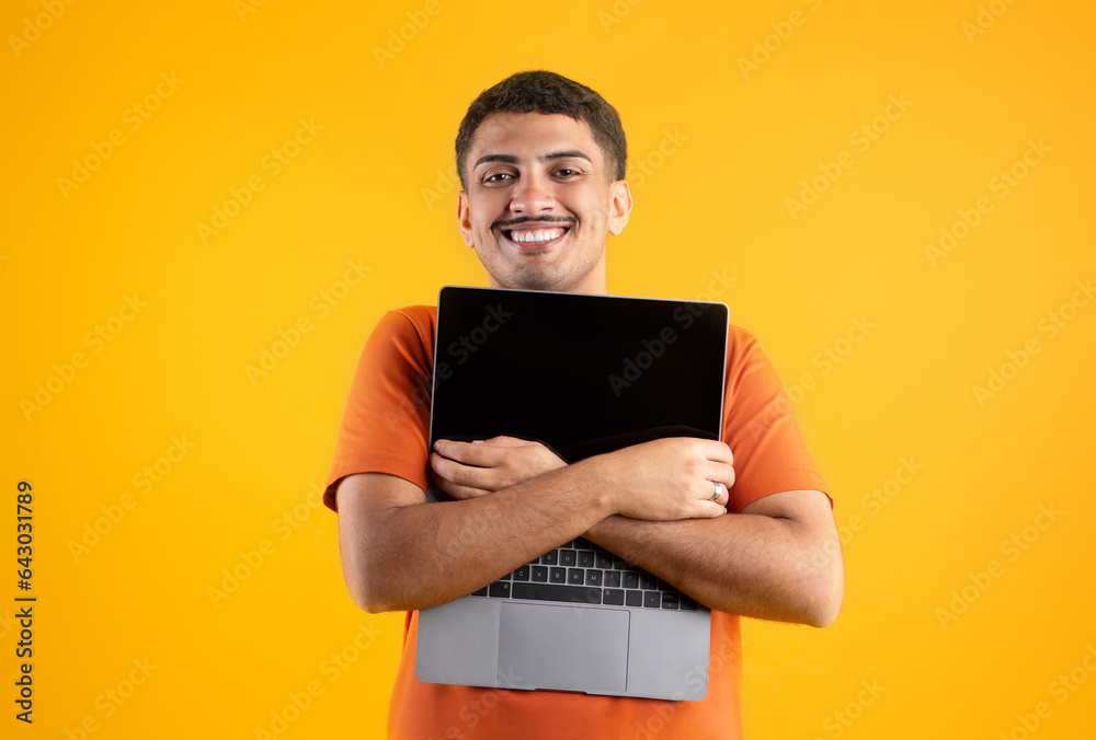 Excited brazilian man embracing modern pc laptop with blank screen and smiling at camera, posing on yellow background