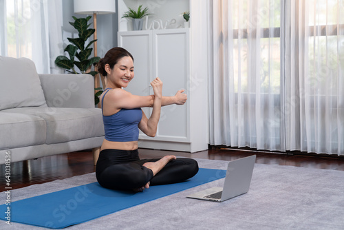 Asian woman in sportswear doing yoga exercise on fitness mat as her home workout training routine. Healthy body care lifestyle woman watching online yoga video on laptop. Vigorous