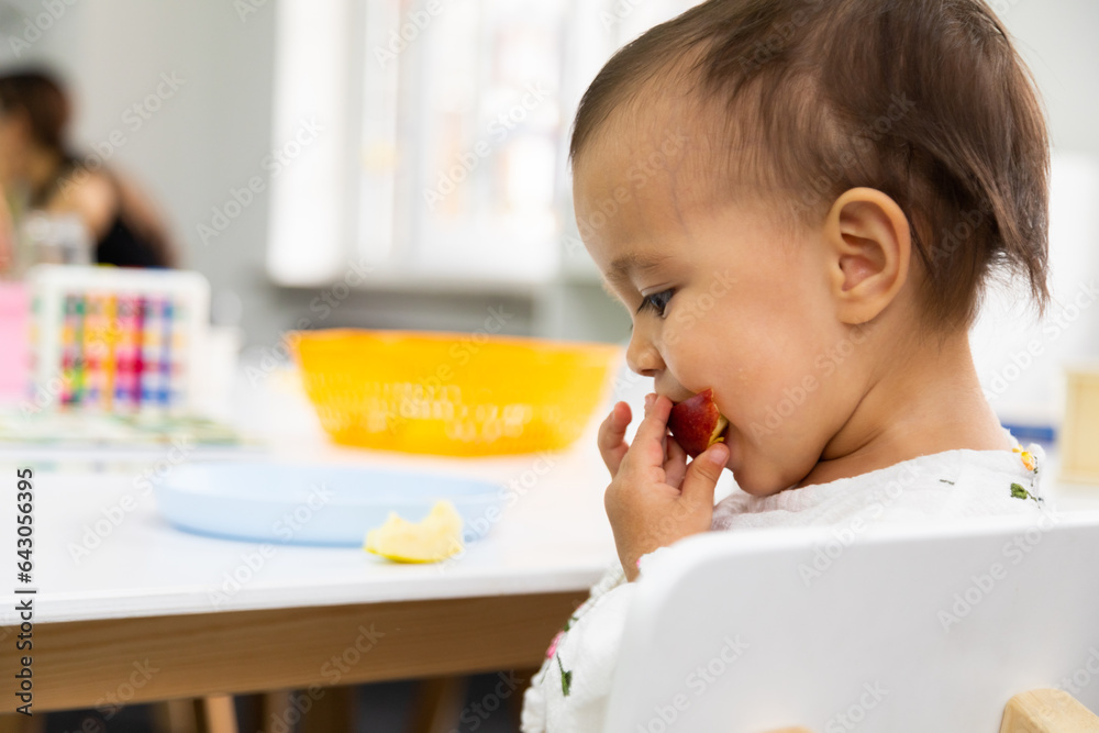 Toddler eating cut apple and sitting by the table in kindergarten - obrazy, fototapety, plakaty 