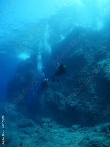  scuba divers exploring around a reef underwater deep blue water big rocks and bubbles ocean scenery  © underocean