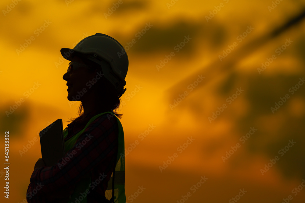 Half-body shot of long-haired female engineer Wear a vest and helmet. Holding a tablet to work on industrial business in a factory Standing in the golden twilight sky scene Half-body shot of long-haired female engineer Wear a vest and helmet. Holding a tablet to work on industrial business in a factory Standing in the golden twilight sky scene