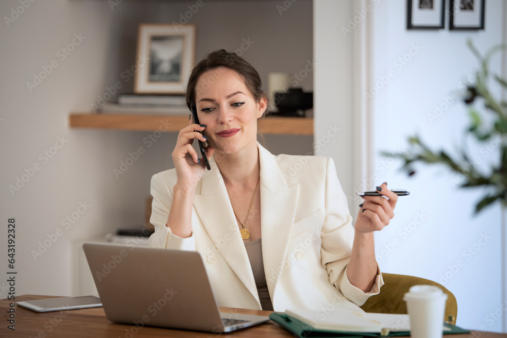 Attractive young businesswoman sitting at desk at home and using smartphone and laptop