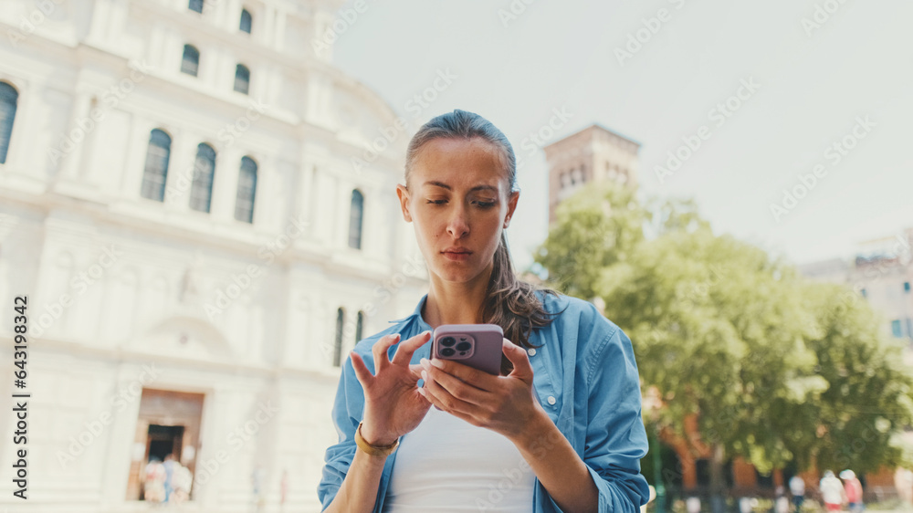Fototapeta premium Beautiful girl with long brown hair, dressed in blue shirt, uses map application on her mobile phone in old town square