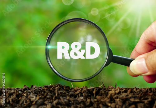 Hands holding magnifying glass focused on R&D text on the soil ground in garden with green natural background.Environmental friendly research and development, Sales, businesses concept.
