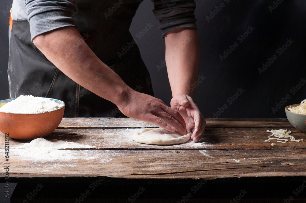 Dark and moody photo of a man hand rolling pizza dough on a vintage wooden counter with bowls of flour, tomato sauce and cheese