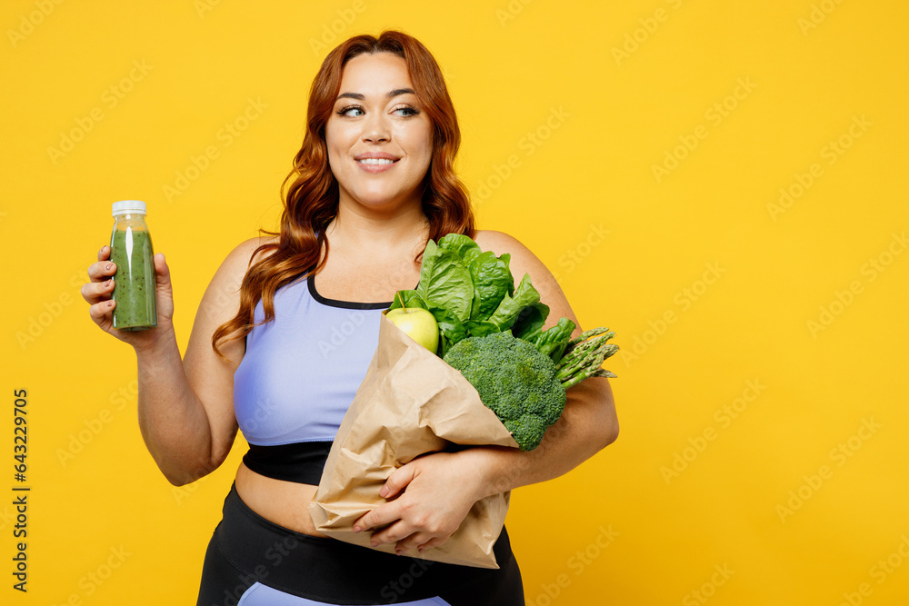 Young chubby plus size big fat fit woman wear blue top warm up train hold smoothie bottle bag with vegetables look aside isolated on plain yellow background studio home gym. Workout sport concept. Young chubby plus size big fat fit woman wear blue top warm up train hold smoothie bottle bag with vegetables look aside isolated on plain yellow background studio home gym. Workout sport concept.