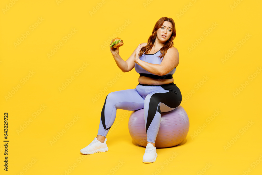 Full body young chubby plus size big fat fit woman wear blue top warm up train sit on fit ball hold burger show stop gesture isolated on plain yellow background studio home gym. Workout sport concept. Full body young chubby plus size big fat fit woman wear blue top warm up train sit on fit ball hold burger show stop gesture isolated on plain yellow background studio home gym. Workout sport concept.