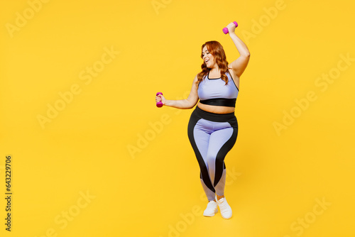 Fototapeta Naklejka Na Ścianę i Meble -  Full body satisfied young chubby plus size big fat fit woman wears blue top warm up training raise up hands with dumbbells isolated on plain yellow background studio home gym. Workout sport concept.