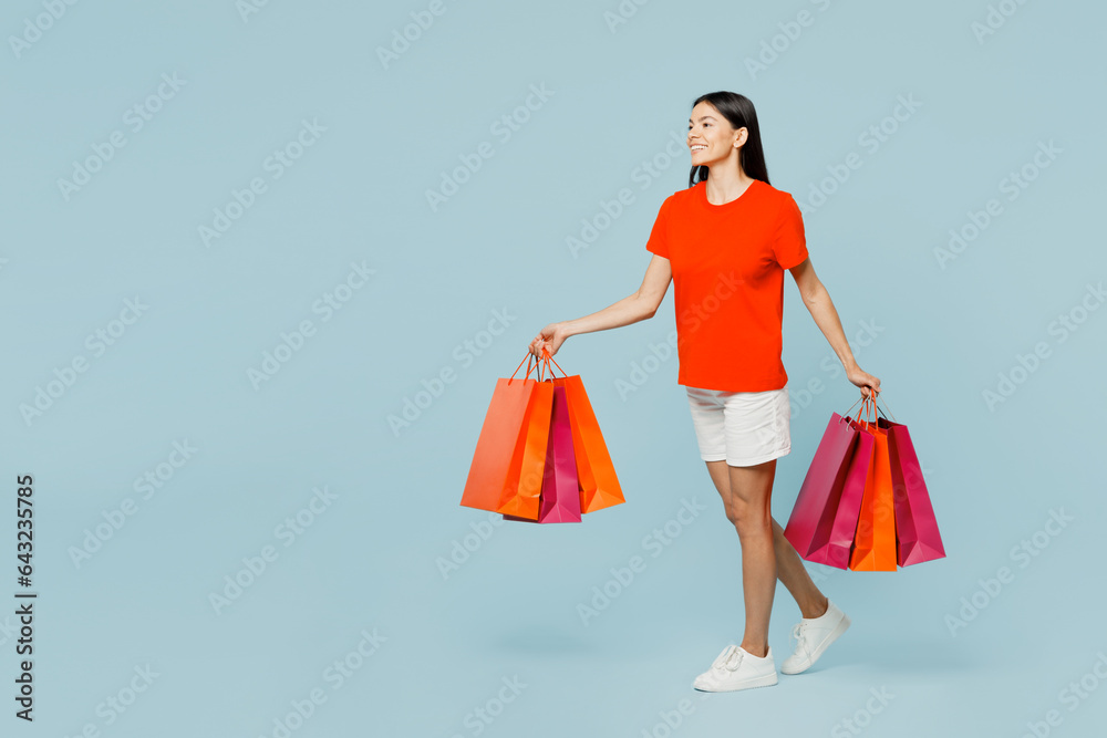 Full body young latin woman wearing orange red t-shirt casual clothes hold paper package bags after shopping go isolated on plain light pastel blue cyan background. Black Friday sale buy day concept. Full body young latin woman wearing orange red t-shirt casual clothes hold paper package bags after shopping go isolated on plain light pastel blue cyan background. Black Friday sale buy day concept.