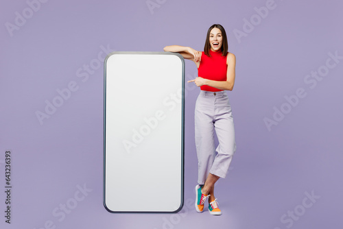 Full body young woman wears red tank shirt casual clothes point finger on big huge blank screen mobile cell phone smartphone with workspace copy space mockup area isolated on plain purple background. photo