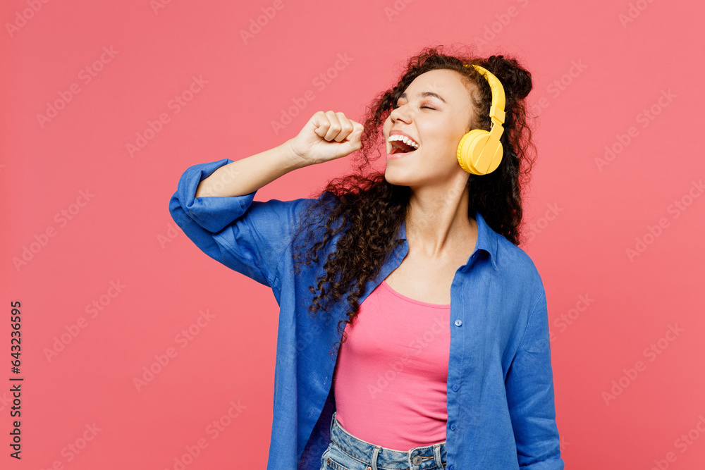 Young woman of African American ethnicity she wear blue shirt casual clothes listen to music in headphones sing song in microphone isolated on plain pastel pink background studio. Lifestyle concept. Young woman of African American ethnicity she wear blue shirt casual clothes listen to music in headphones sing song in microphone isolated on plain pastel pink background studio. Lifestyle concept.