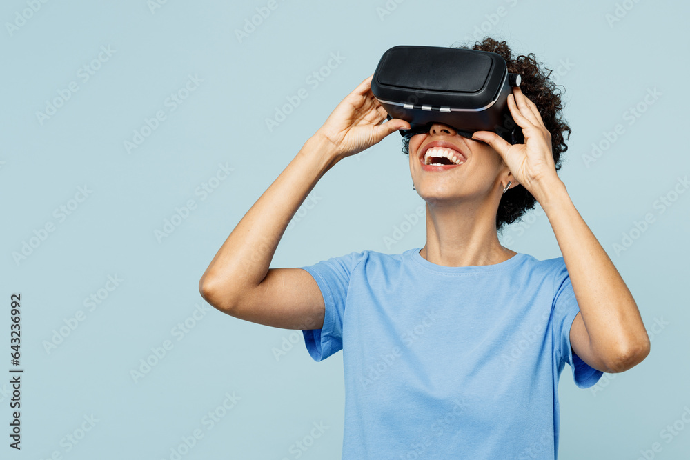 Young amazed impressed fun cool woman of African American ethnicity wear t-shirt casual clothes watching in vr headset pc gadget isolated on plain pastel light blue cyan background. Lifestyle concept. Young amazed impressed fun cool woman of African American ethnicity wear t-shirt casual clothes watching in vr headset pc gadget isolated on plain pastel light blue cyan background. Lifestyle concept.