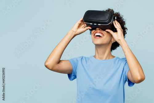Young amazed impressed fun cool woman of African American ethnicity wear t-shirt casual clothes watching in vr headset pc gadget isolated on plain pastel light blue cyan background. Lifestyle concept. © ViDi Studio