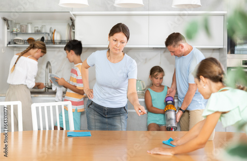 Mom tells daughter to wipe kitchen table thoroughly, husband and younger children do kitchen chores in background