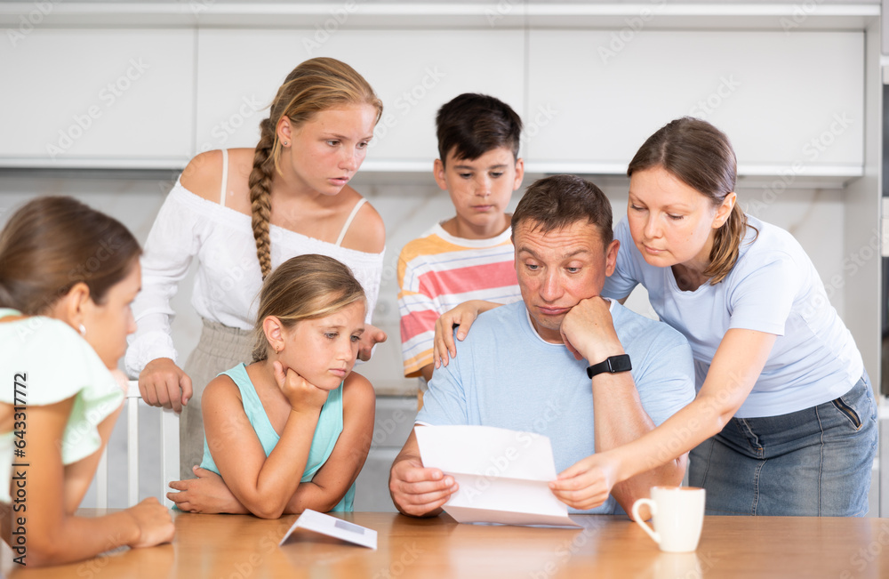 Fototapeta premium Large family is standing next to confused man sitting near kitchen table. Man reads letter and is upset by bad news from collector, unexpected test results, increase in loan rate