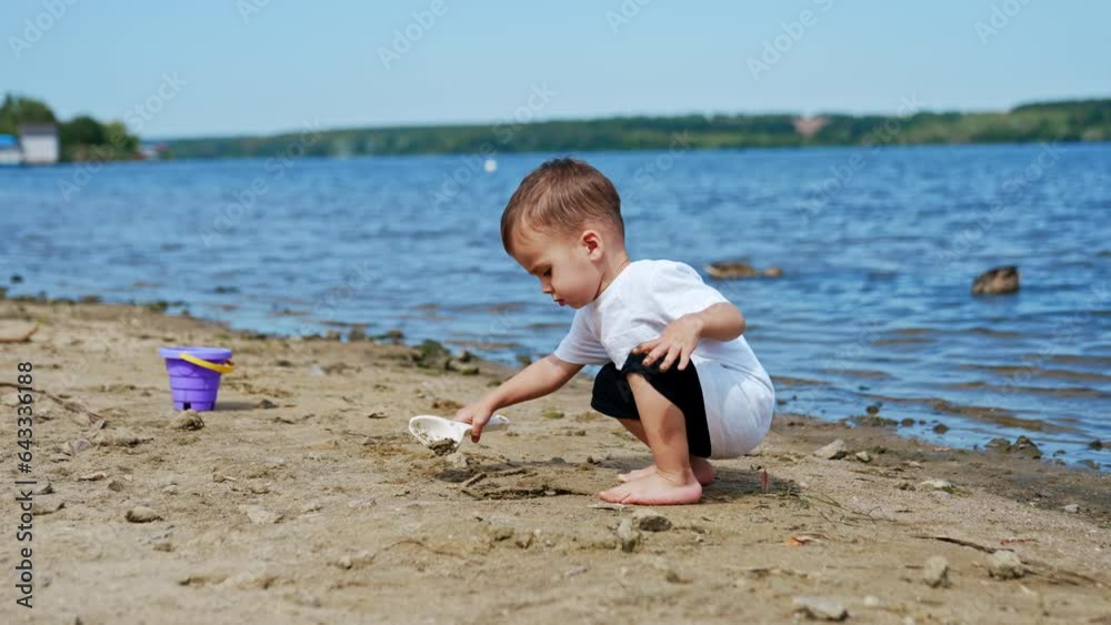 Little Caucasian boy in white t-shirt digging sand on the beach. Toddler working with shovel on the shore. Little Caucasian boy in white t-shirt digging sand on the beach. Toddler working with shovel on the shore.
