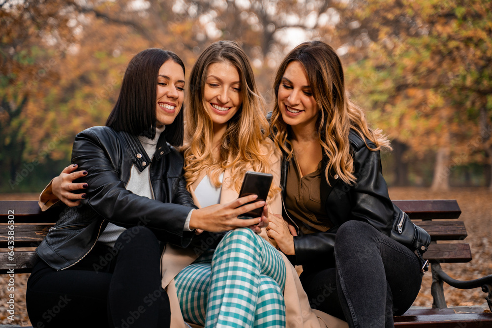 Three young fashionable young woman sitting on park bench and looking at smartphone together, watching video clips Three young fashionable young woman sitting on park bench and looking at smartphone together, watching video clips