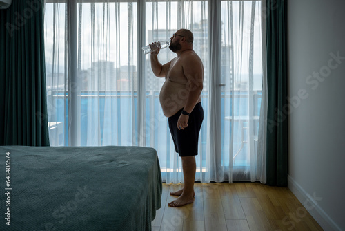 Thirsty overweight man feeling dehydration drinking water in hotel room in hot humid weather standing near window. Fat guy with naked torso suffers from heat, heat stroke, overheating, excess weight. photo
