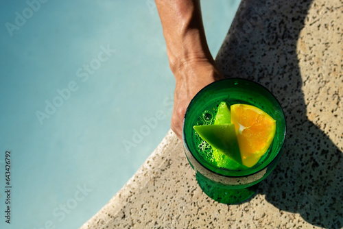 Close em um drinque refrescante com rodelas de limão e laranja sendo segurado por mão de mulher na beira da piscina em dia de sol. 