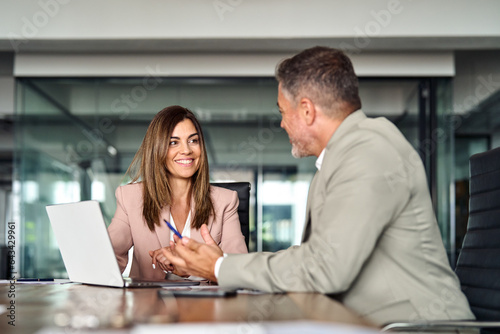 Happy professional mature Latin businessman and businesswoman executive colleagues wearing suits sitting at table having partnership business discussion using laptop at corporate meeting in office.