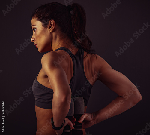 Female muscular doing stretching and strength workout on the shoulders, blades and arms in sport bra holding dumbbells back behind herself standing on grey studio background with empty space.