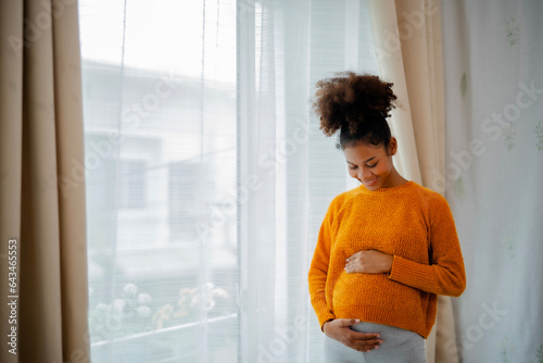 African American pregnancy woman in yellow sweater standing beside glasses door, embrassing her belly with smile of care and love moment for baby. photo