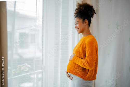 African American pregnancy woman in yellow sweater standing beside glasses door, embrassing her belly, looking out of window with smile of love and happiness. photo