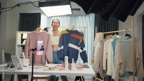 Young Asia lady seller look at phone camera live stream use laptop answer follower comment introduce sweater new collection new arrival on her chanel at studio. Woman fashion online shop concept.