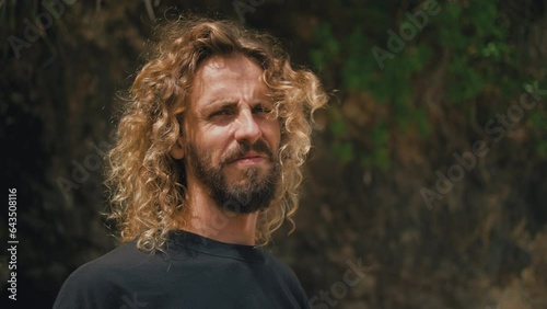 Blinking in sunlight curly surfer with long hair on idyllic beach. Male hippie traveler on seaside. Caucasian man with beard on ocean shore. Enjoying summer vacation in tropical paradise. Travel.