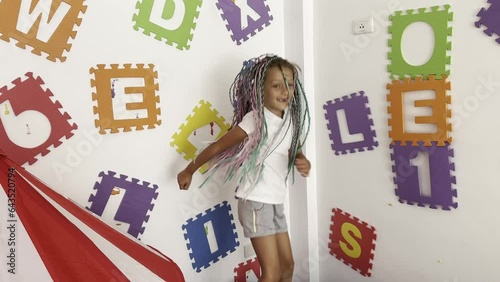 Happy little girl in white T-shirt and many colored braids on head jumps on children's trampoline in Gymboree.  Funny holiday and new childhood experiences.