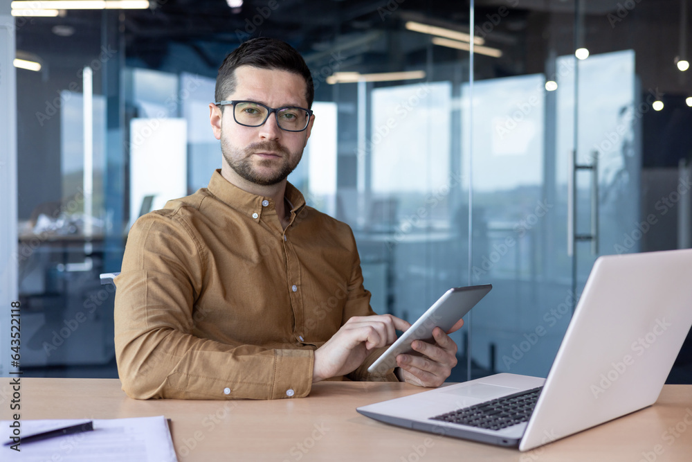 Portrait of a young male office worker sitting at a desk, holding a tablet and looking seriously at the camera Portrait of a young male office worker sitting at a desk, holding a tablet and looking seriously at the camera