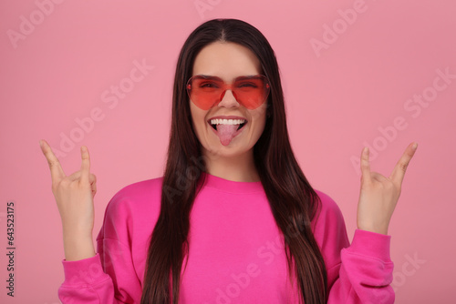 Happy young woman with heart shaped glasses showing her tongue and making rock gesture on pink background